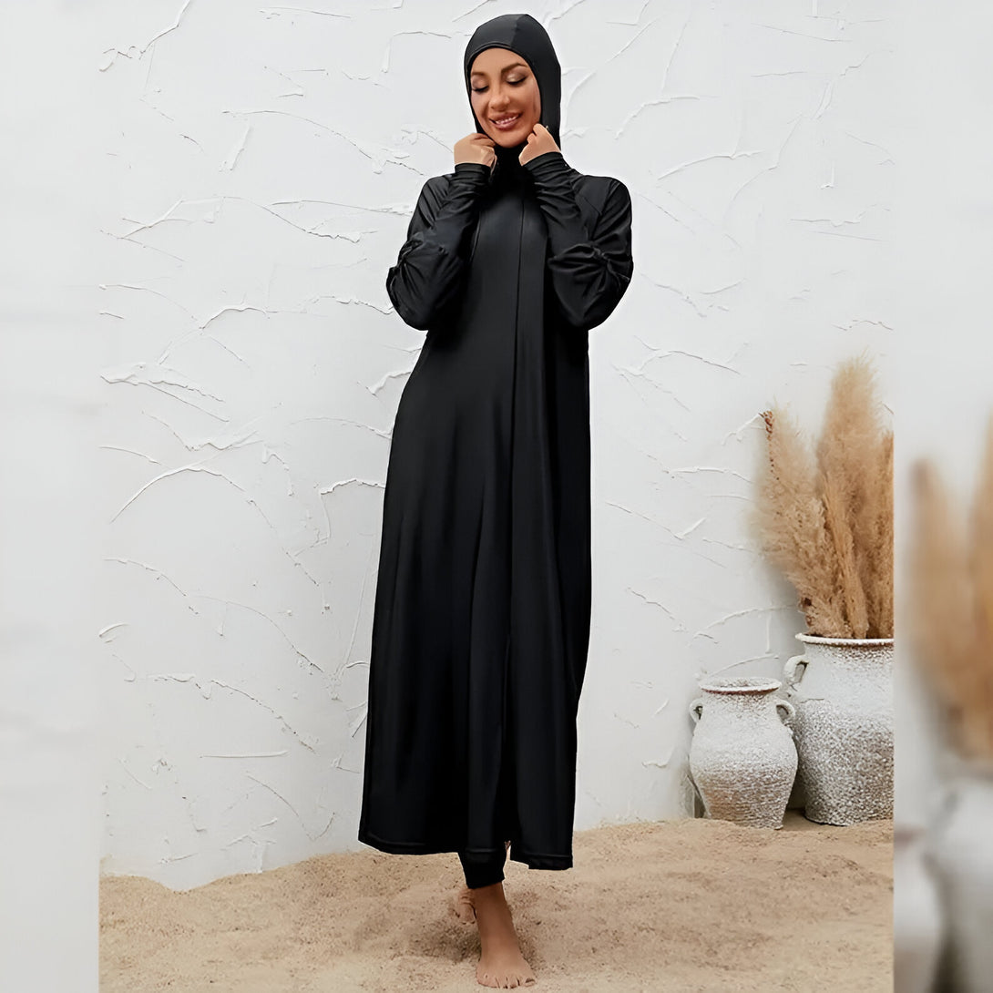 Une femme portant un hijab noir et une longue robe de bain noire, debout sur le sable. Vêtements de bain musulmans pour femmes.