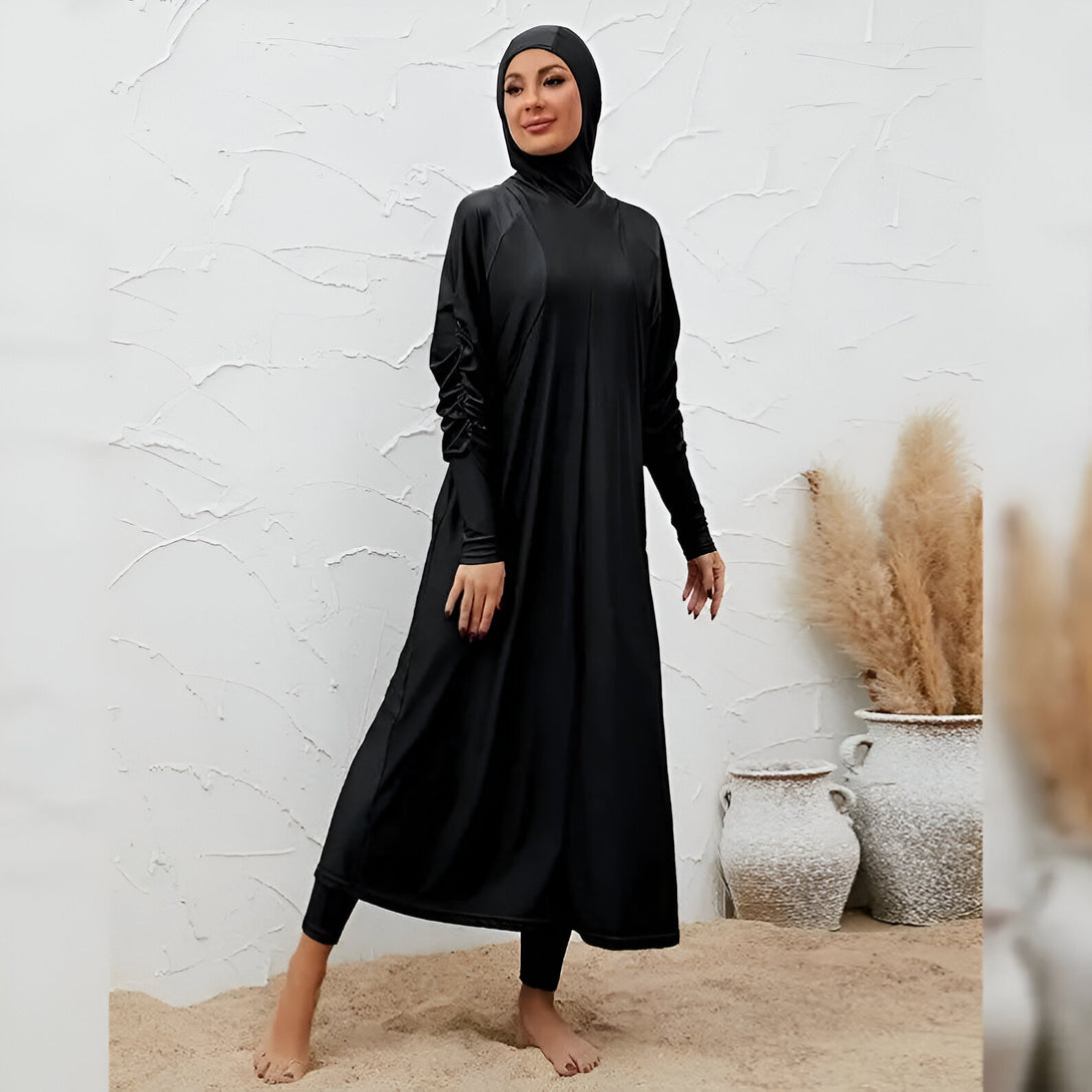 Une femme portant un maillot de bain musulman noir avec une longue robe et un hijab assorti, debout sur le sable.