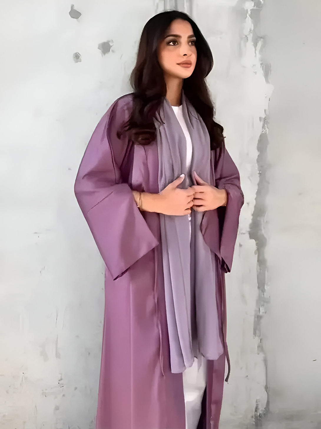 Une femme élégante porte une abaya violette et un hijab assorti. Elle est debout devant un mur gris clair. Cette abaya est parfaite pour les occasions spéciales ou le quotidien.