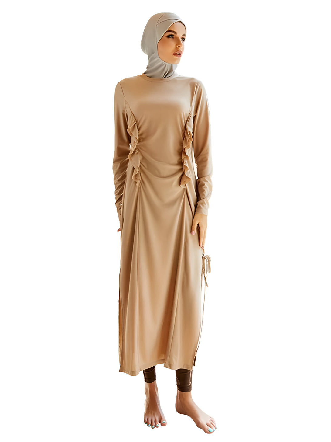 Une femme porte une robe longue beige à manches longues avec une fente sur les côtés et un hijab gris. La robe est cintrée à la taille et a des volants sur les côtés.
