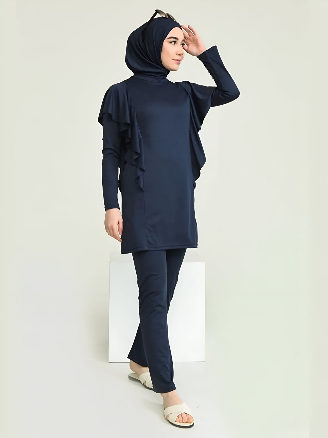 Une femme portant un maillot de bain musulman bleu marine avec des volants. Le maillot de bain comprend un haut à manches longues et un pantalon long. La femme porte également un hijab bleu marine et des sandales blanches.
