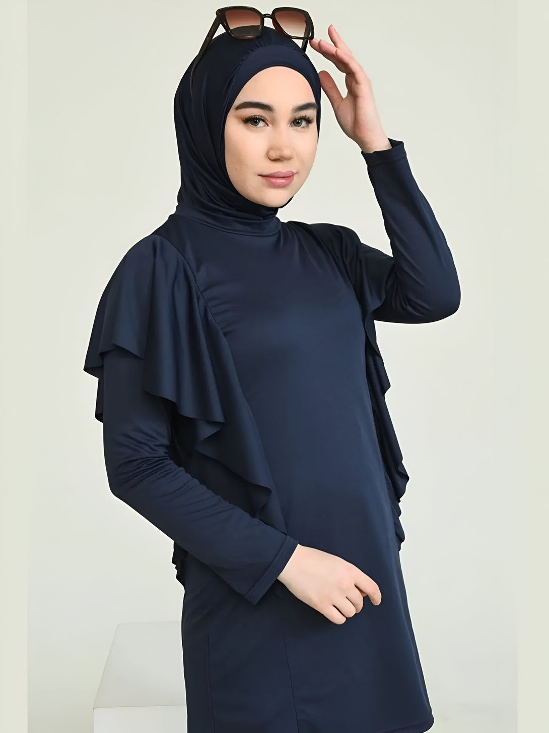 Une femme portant une robe bleu marine à manches longues et un hijab assorti. Elle porte également des lunettes de soleil. La robe a des volants sur les épaules.