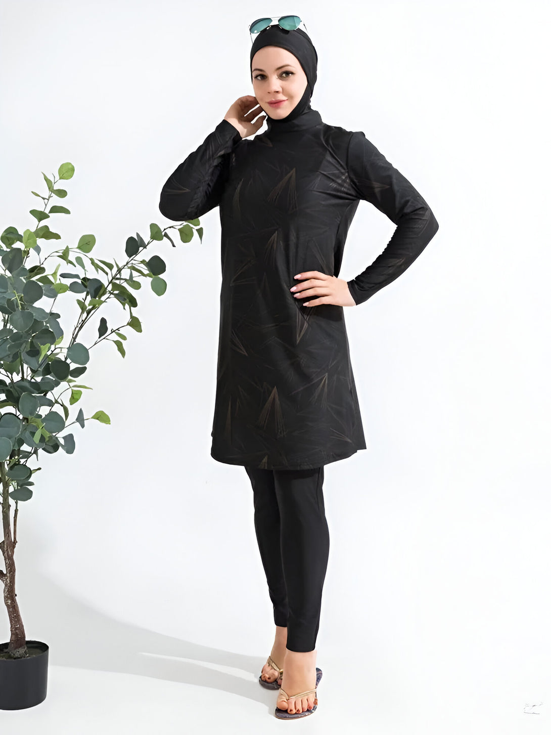 Une femme portant un maillot de bain musulman noir avec un imprimé géométrique doré. Le maillot de bain est à manches longues et comprend un pantalon long. Elle porte également un hijab noir et des lunettes de soleil.
