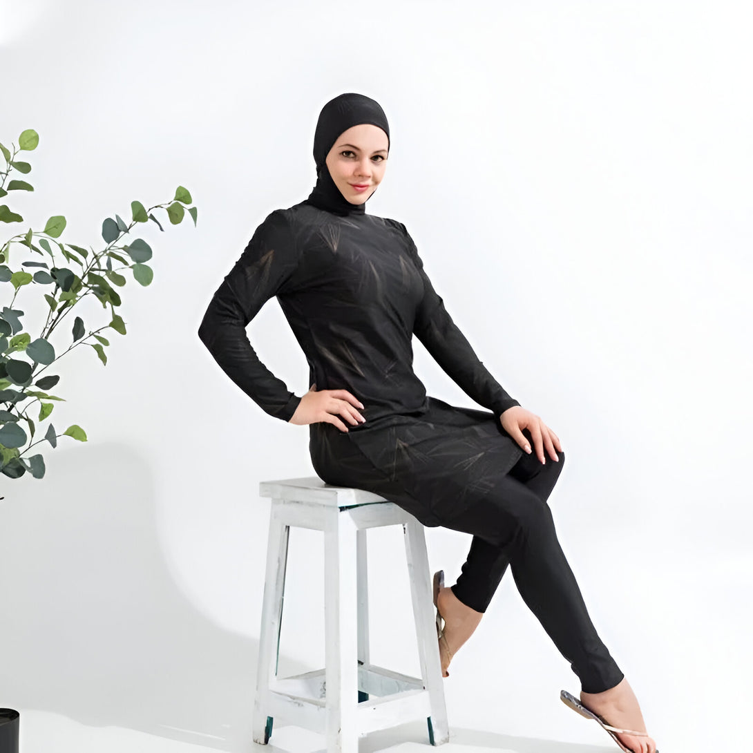 Une femme portant un maillot de bain musulman noir à imprimé or, assis sur un tabouret blanc.  Elle porte un hijab noir.