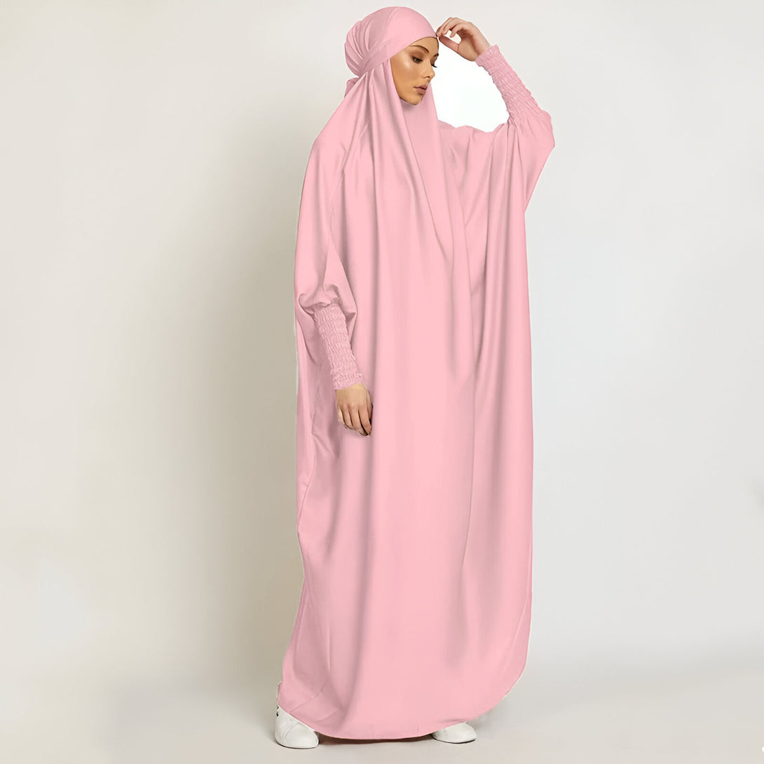 Une femme portant une longue robe rose avec des manches larges et un hijab assorti. La robe est ample et confortable, idéale pour la prière ou les occasions spéciales.