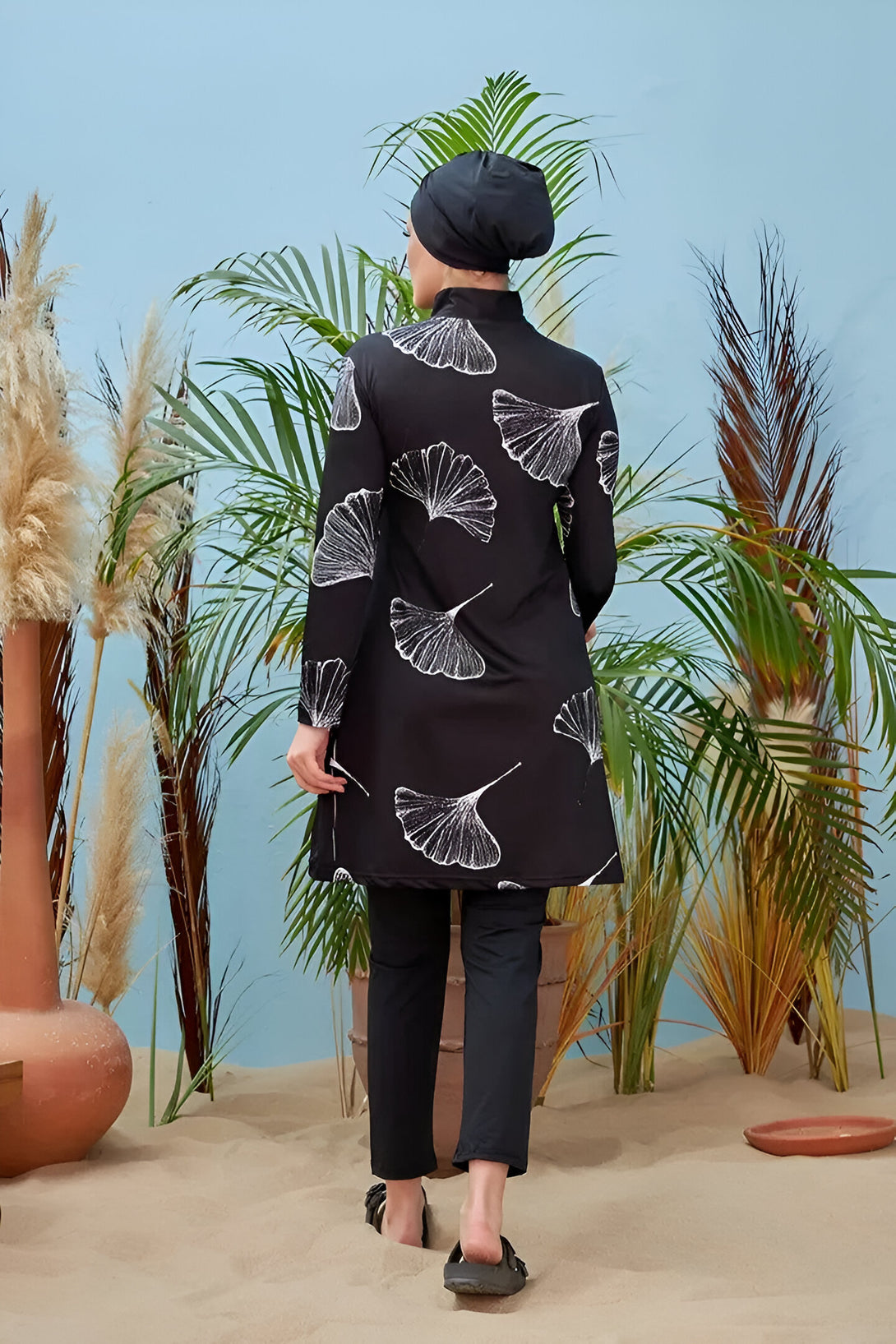 Une femme portant un maillot de bain musulman noir avec un imprimé de feuilles blanches, un hijab noir et des sandales noires marche sur le sable.