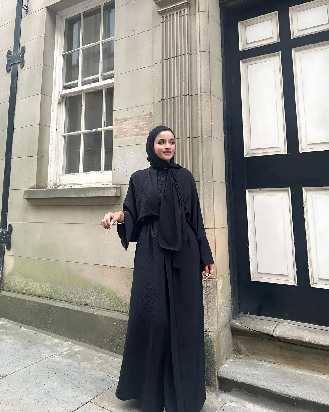 Une femme portant une abaya noire élégante et un hijab assorti, posant devant un bâtiment en pierre.  La robe est à la fois modeste et élégante, idéale pour les occasions spéciales ou le quotidien.  Découvrez notre collection d'abayas et trouvez votre style.