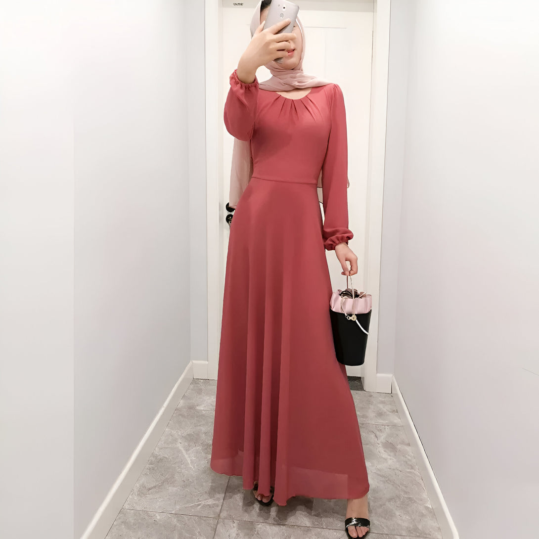 Une femme portant une robe longue rose à manches longues et un hijab rose. Elle tient un sac à main noir et prend une photo d'elle-même dans un miroir.