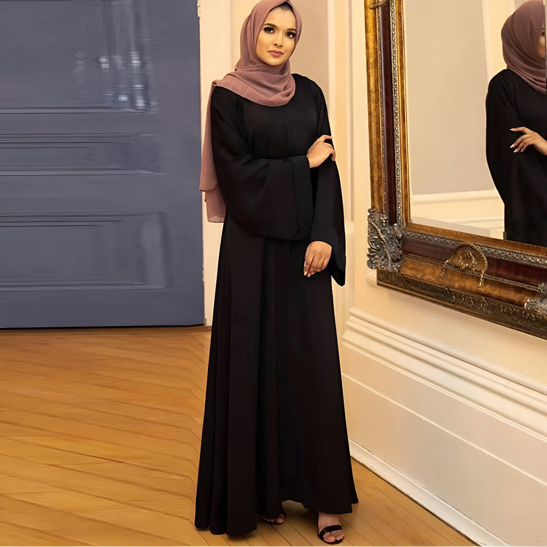Une femme portant une abaya noire élégante avec une large ceinture et un hijab rose devant un miroir.