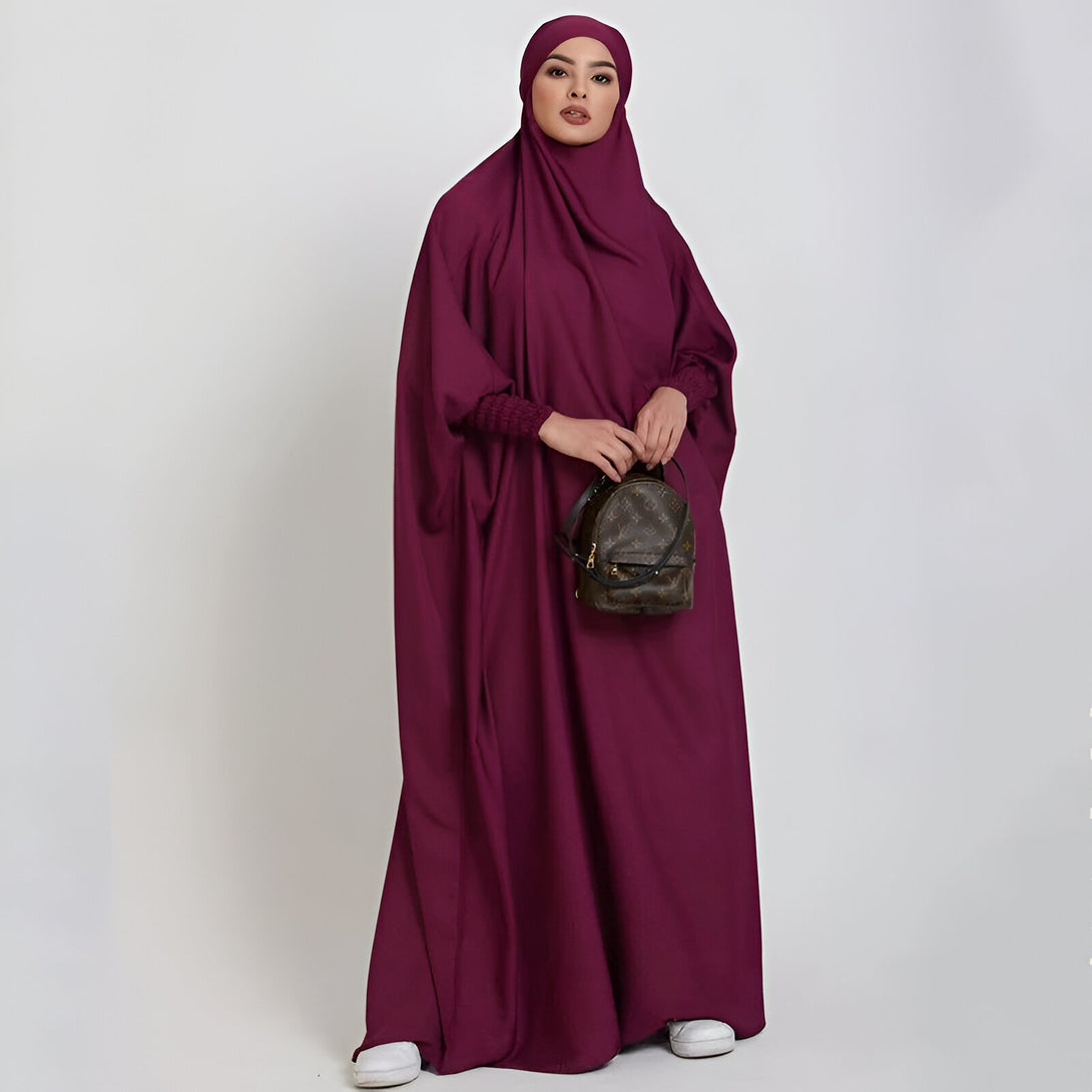 Une femme portant une abaya longue et ample de couleur bordeaux, avec un hijab assorti. Elle porte un petit sac à dos marron sur l'épaule.