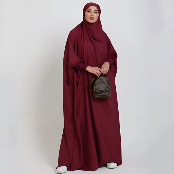 Une femme portant une abaya bordeaux élégante avec un hijab assorti, tenant un sac à dos brun.