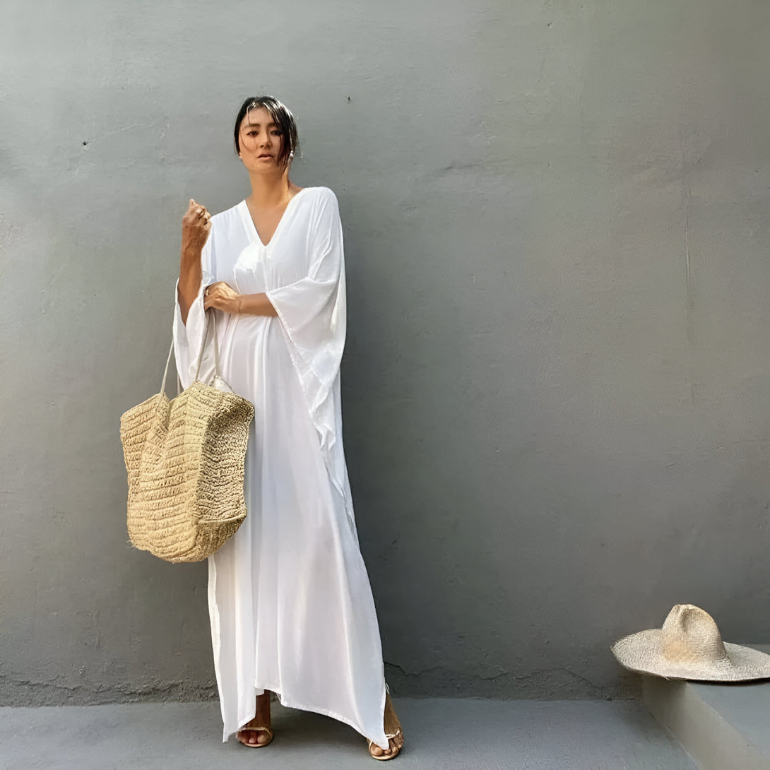 Une femme porte une longue robe blanche ample et un sac à main en paille. Elle est debout devant un mur gris.
