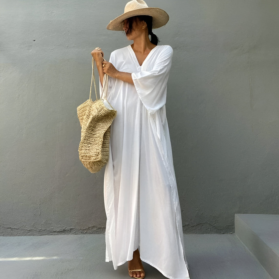 Une femme porte une longue robe blanche ample avec un décolleté en V et des manches amples. Elle porte un chapeau de paille et un sac en paille. La robe est parfaite pour les occasions décontractées ou formelles.