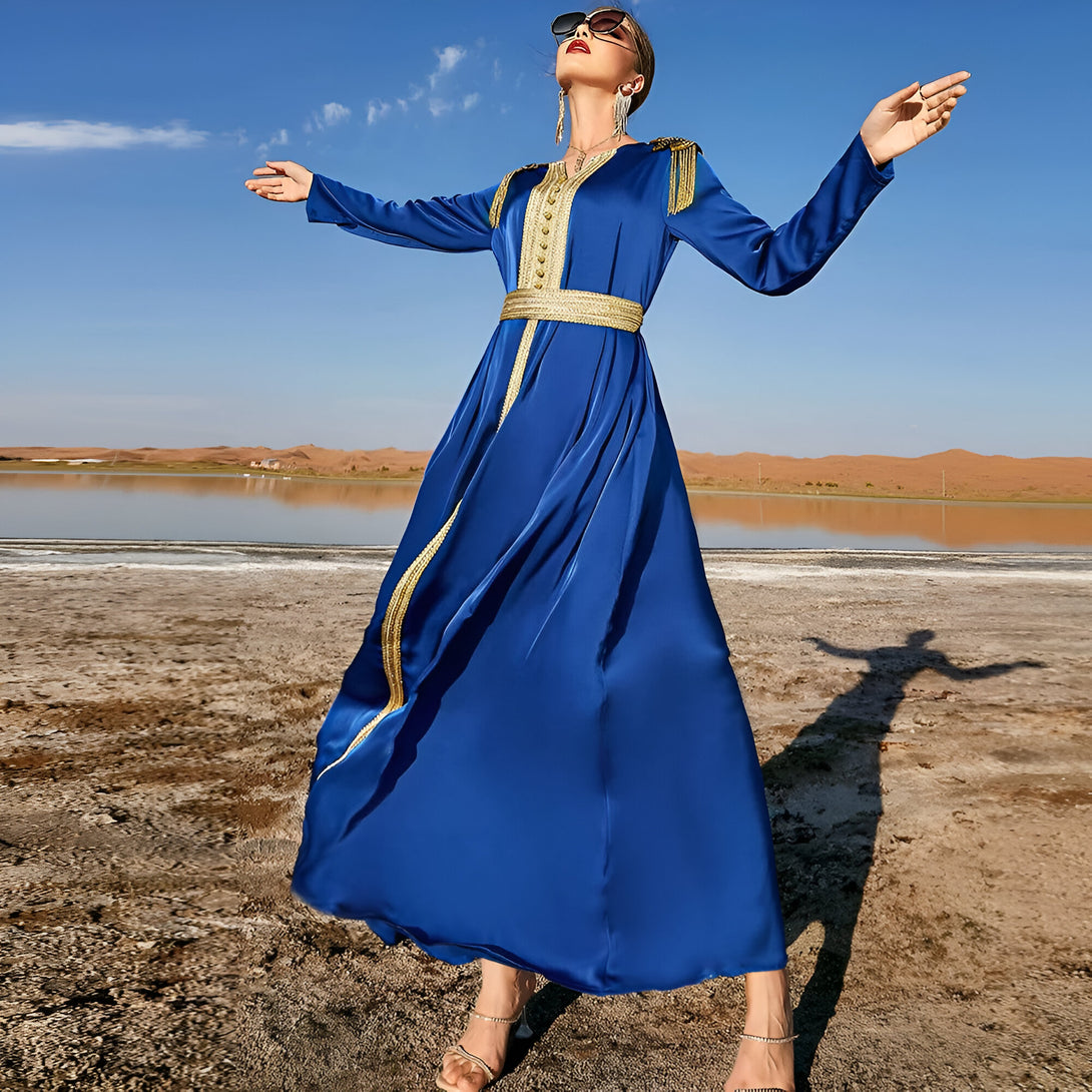 Une femme porte une robe longue bleue avec des détails dorés, debout sur une plage désertique. La robe est élégante et a un design moderne, parfait pour les occasions spéciales ou les événements formels.