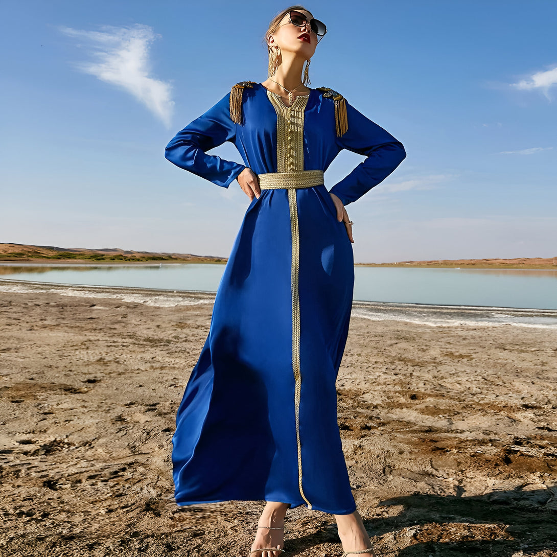 Une femme portant une longue robe bleue avec des détails dorés et une ceinture. Elle porte des lunettes de soleil et se tient sur un fond de plage.
