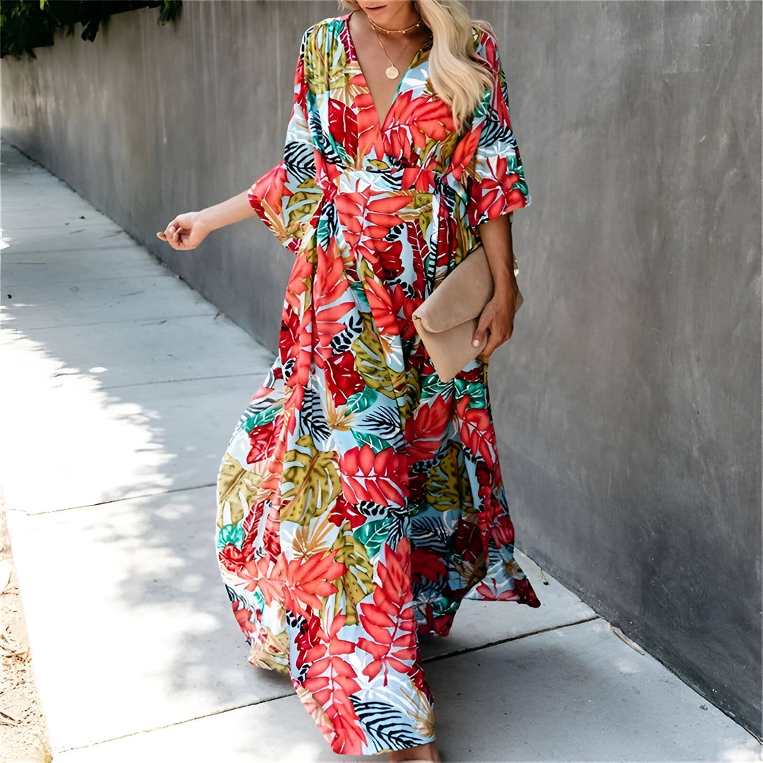 Une femme portant une longue robe à fleurs avec une encolure en V et des manches 3/4. La robe est à imprimé floral avec des couleurs vives. La femme porte également un sac à main beige.