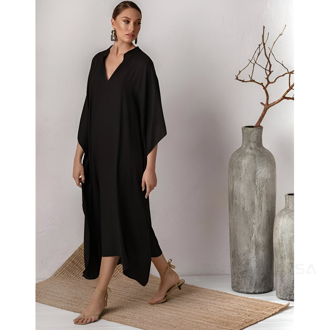 Une femme porte une longue robe noire ample avec un col en V. La robe a des manches larges. La femme porte des sandales à talons et des boucles d'oreilles. Elle se tient debout sur un tapis en jute devant un vase en céramique gris.