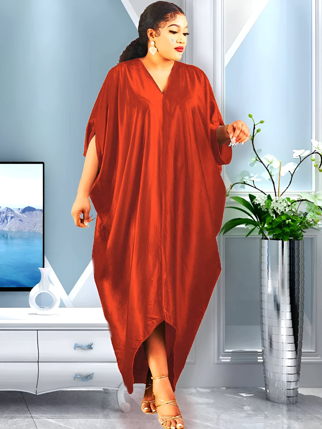 Une femme porte une longue robe rouge ample et élégante avec un décolleté en V. Elle a les cheveux noirs et porte des sandales dorées. La robe est parfaite pour les occasions spéciales et les sorties décontractées.